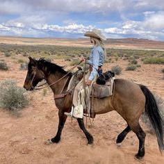 a man riding on the back of a brown horse across a dirt field with a dog