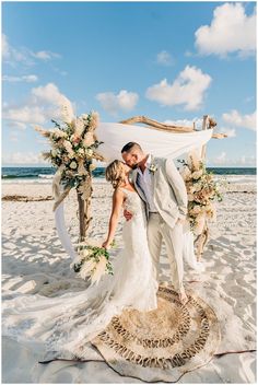 a bride and groom kissing on the beach in front of their wedding arch with flowers