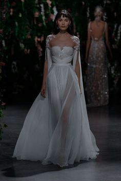 a model walks down the runway in a white gown with sheer sleeves and an embellished cape