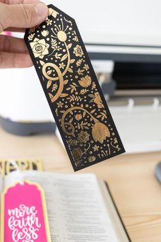 a person is holding up a bookmark that has gold designs and flowers on it