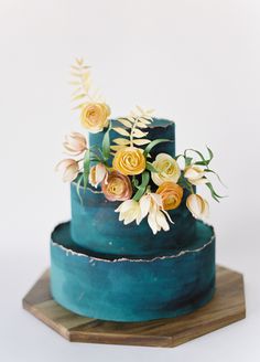 a three tiered blue cake with flowers on the top and bottom, sitting on a wooden stand