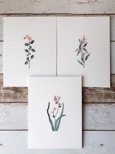 three cards with watercolor flowers on them sitting on a wooden table next to each other