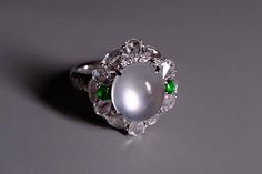 Fine jade ring set with high quality 'glassy' white (main) and green (small) jadeite cabochons and accented with diamonds in 18k. The jade has a very fine grain crystal and is noted for its transparency. In jadeite, this '玻璃种' or glassy type is distinguished from mere icy or translucent jade. Glassy jadeite has the topmost clarity in terms of transparency. Specifically, these is icy white jade that displays an optical phenomenon known in the jade trade as '起莹光' or luminescence. Luminescence appears as a silvery white light moving through the jade cabochon. The phenomenon is not the reflection of light/lamp but pushed from within the stone. If present, this phenomenon is mostly observed in top quality icy white jadeite, and less commonly in icy green jadeite. "Every jade is unique": this di White Multi-stone Moonstone Ring For Formal Occasions, Formal White Multi-stone Moonstone Ring, Elegant Green Cabochon Moonstone Ring, Elegant White Jade Ring, Optical Phenomena, White Jade, Jade Ring, Light Reflection, Light Lamp