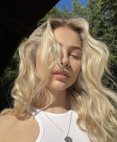 a woman with long blonde hair wearing a white tank top and silver pendants on her necklace