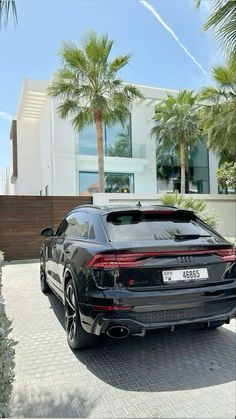 the rear end of a black car parked in front of a large white house with palm trees