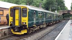 a green and yellow train traveling down tracks next to a loading platform at a train station