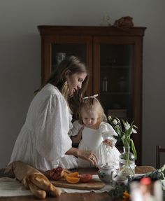 a woman and child sitting at a table in front of flowers, bread and fruit