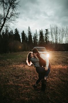 a man and woman kissing in front of a car on a field with trees behind them