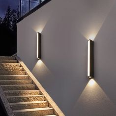 three lights on the side of a white wall next to stairs and windows at night