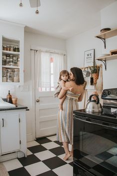 a woman holding a child in her arms while standing next to an oven and stove top