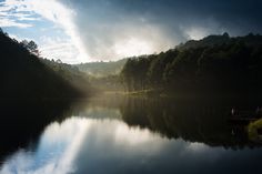 a body of water surrounded by trees under a cloudy sky with sun shining through the clouds