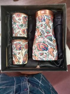 two matching flasks are in a box with leather sleeves and floral designs on them