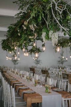 a long table with white chairs and greenery hanging from it's ceiling is set for an event