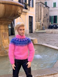 a barbie doll wearing a pink sweater and black pants standing in front of a fountain