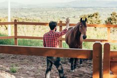 a man standing next to a brown horse behind a wooden fence on top of a dirt field