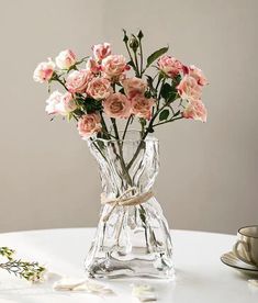 a glass vase filled with pink flowers on top of a white table next to a cup and saucer