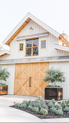 a large white barn with wooden doors and windows on the side of it, surrounded by greenery