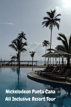 an outdoor pool with lounge chairs and umbrellas next to the ocean in front of palm trees