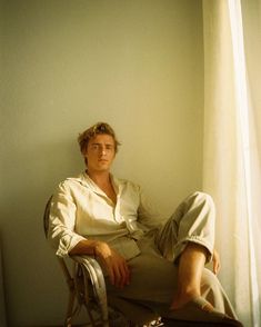 a man sitting on top of a chair next to a white wall with a window behind him