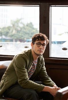 a young man sitting in front of a window wearing glasses and a green jacket with striped shirt