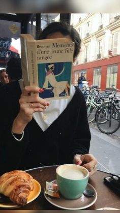 a woman sitting at a table reading a book and eating croissants in front of her