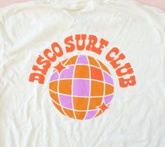 This fun and retro t-shirt features a bold "DISCO SURF CLUB" graphic with a groovy orange and purple disco ball at the center. Its nostalgic, laid-back design blends 1970s disco vibes with the carefree energy of surf culture, making it the perfect statement piece for any casual outfit. Fit: Classic fit: Comfortable and slightly loose for easy all-day wear. Standard length: Hits at the hips, perfect for tucking into jeans or pairing with shorts. Short sleeves: Timeless and versatile, great for la Purple Disco Ball, Purple Disco, Disco Vibes, 1970s Disco, Surf Club, Surf Culture, Striped Bags, Fashion Bottoms, Turkish Towels Beach