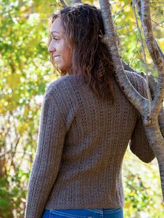 a woman standing in front of a tree with her back to the camera, wearing a brown sweater