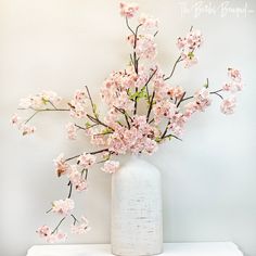 a white vase filled with pink flowers sitting on top of a table next to a wall