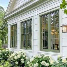 a house with green windows and white flowers in the front yard, next to some bushes