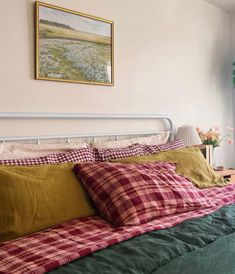 a bed with plaid sheets and pillows in a bedroom next to a window, framed painting on the wall