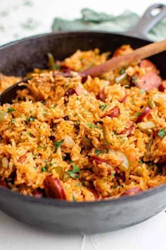 a skillet filled with rice and vegetables on top of a white tablecloth next to a wooden spoon