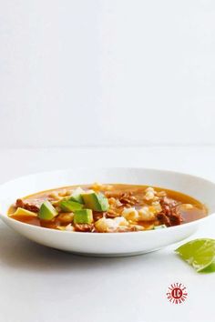 A white bowl filled with posole soup, garnished with diced avocado and surrounded by a lime wedge on a clean white background.