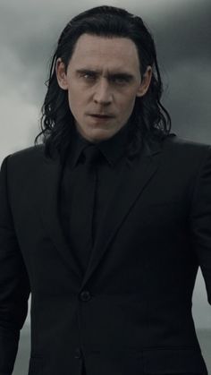 a man with long hair wearing a black suit and standing in front of dark clouds