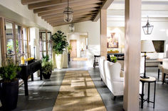 a living room filled with white furniture and lots of windows next to a hallway covered in potted plants