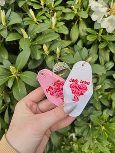 someone holding two small tags in front of some green plants and bushes, one with the words self love country club written on it