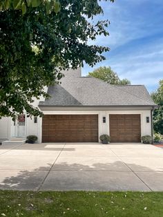 a white house with two brown garage doors on the front and side of it, surrounded by trees