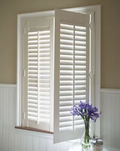 a vase filled with purple flowers sitting next to a window covered in white shutters