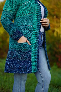 a woman wearing a green and blue crocheted cardigan standing in the grass