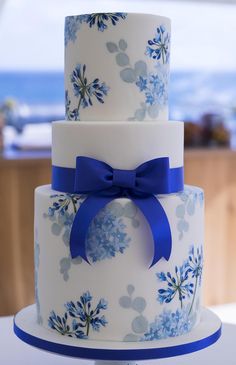 a three tiered cake with blue flowers and a bow on the top is sitting on a table