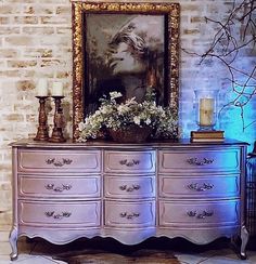 an ornate dresser with flowers on it in front of a brick wall and a painting