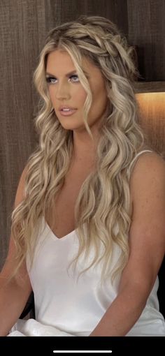 a woman with long blonde hair sitting in a chair wearing a white dress and braiding her hair