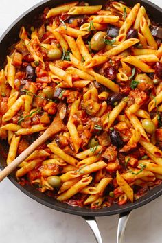 a skillet filled with pasta, olives and meat in tomato sauce next to a wooden spoon