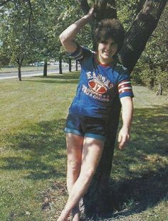 a young man standing next to a tree on top of a lush green park ground