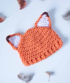 an orange crocheted fox hat laying on top of a white table next to leaves