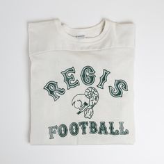 Vintage '69-'81 era Champion "Regis Rams" Lastone print Football Game Jersey. Made of durable cotton/nylon Durene material, this jersey sports larger "TV numbers" and nameplant graphics on the back along with authentic signs of wear. Sizing Fits true to size or slightly larger (these were to have pads underneath). Provided below are additional measurements to aid in gauging fit. Measurements 42" chest / 21" pit to pit 32" long 25" shoulder to shoulder 19" sleeve length Condition Garment is in go Retro Cotton Tops With Team Name, Retro Tops For Football Season Sports Events, Retro Sweatshirt For Sports Events, Retro Graphic Print Sweatshirt For Sports, Retro Tops With Graphic Print For Football Season, Vintage Sports Tops For Football Season, Retro Graphic Print Top For Football Season, Vintage Cotton Sweatshirt For Sports Events, Vintage Relaxed Fit T-shirt For Game Day