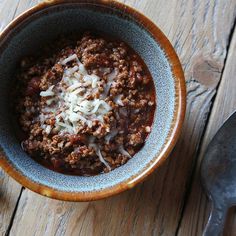 a bowl filled with chili and cheese next to a spoon on top of a wooden table