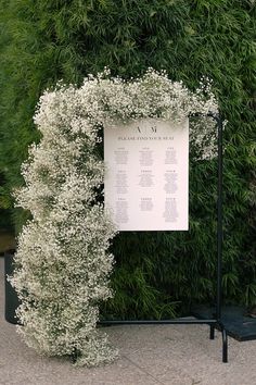 a white sign sitting on top of a lush green plant covered wall next to a sidewalk