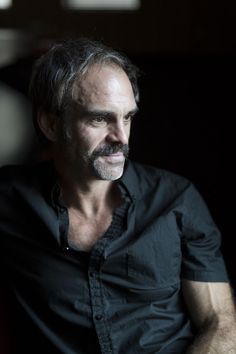 a man in black shirt sitting down with his hand on his chest and looking off to the side