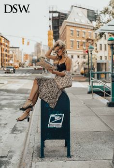 a woman sitting on top of a mailbox reading a paper while wearing high heels