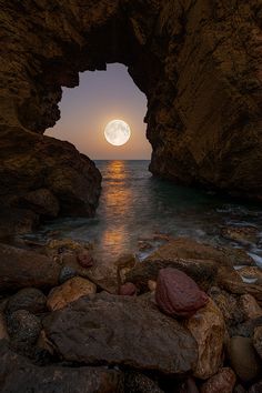 the moon is setting over the ocean with rocks in front of it and an arch leading into